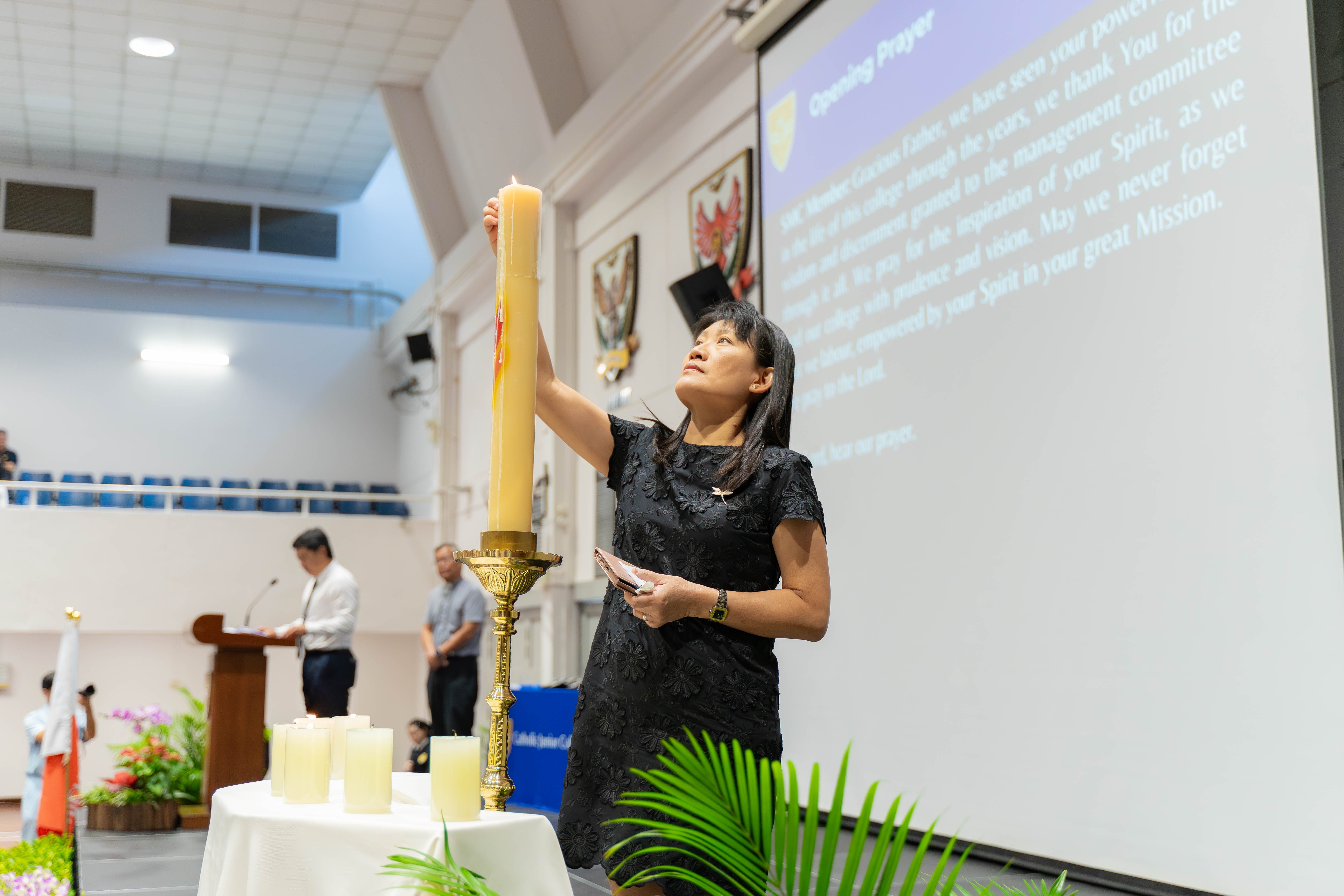 Woman in black dress lighting a tall candle on a brass holder at a formal ceremony with a prayer projected on screen.